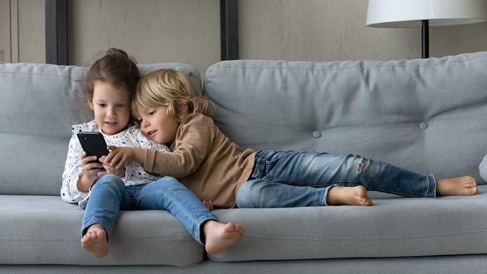 Photo of two young children lounging on a grey couch while playing on a phone.